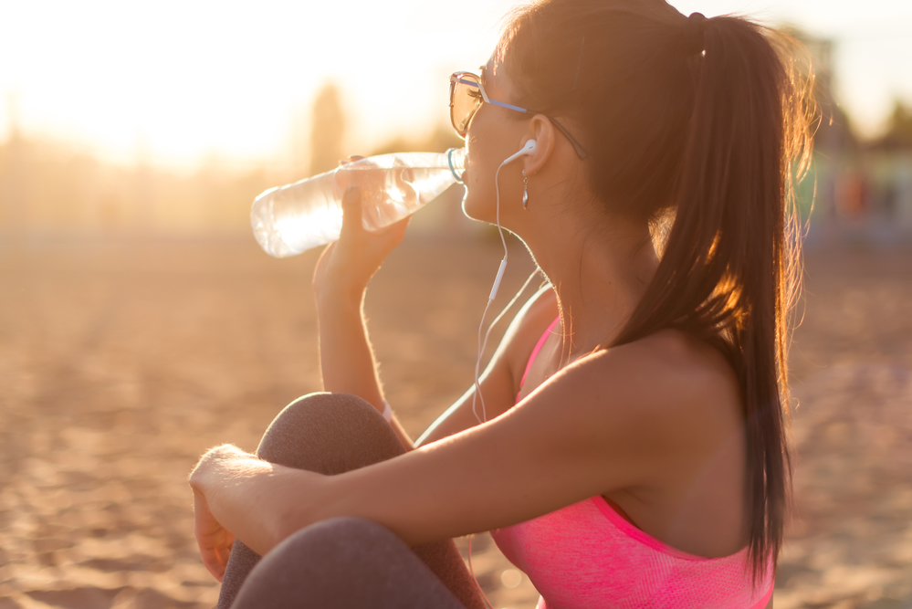 Beautiful fitness athlete woman drinking water after work out exercising on sunset evening summer in beach outdoor portrait.